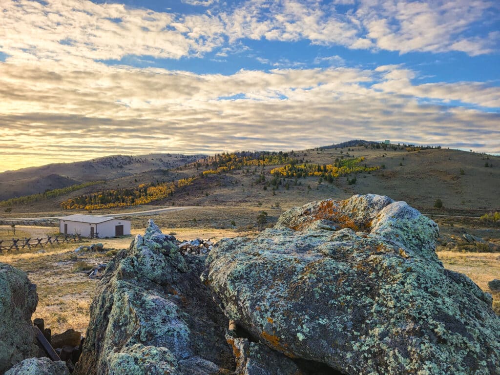 Large moss-covered rocks in the foreground overlook recreational land with rolling hills, yellow-leaved trees, a small building, and a dramatic sky filled with scattered clouds.