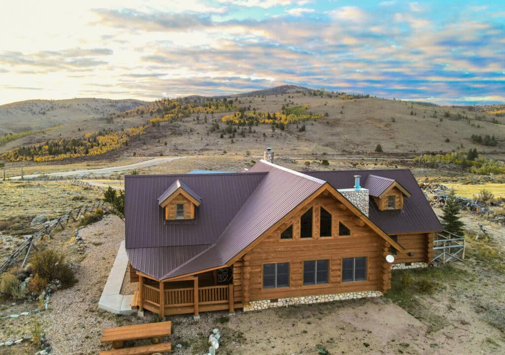 A rustic log cabin with a metal roof sits on recreational land in an open, hilly landscape under a partly cloudy sky, surrounded by sparse trees and dry grass, with rolling hills in the background.