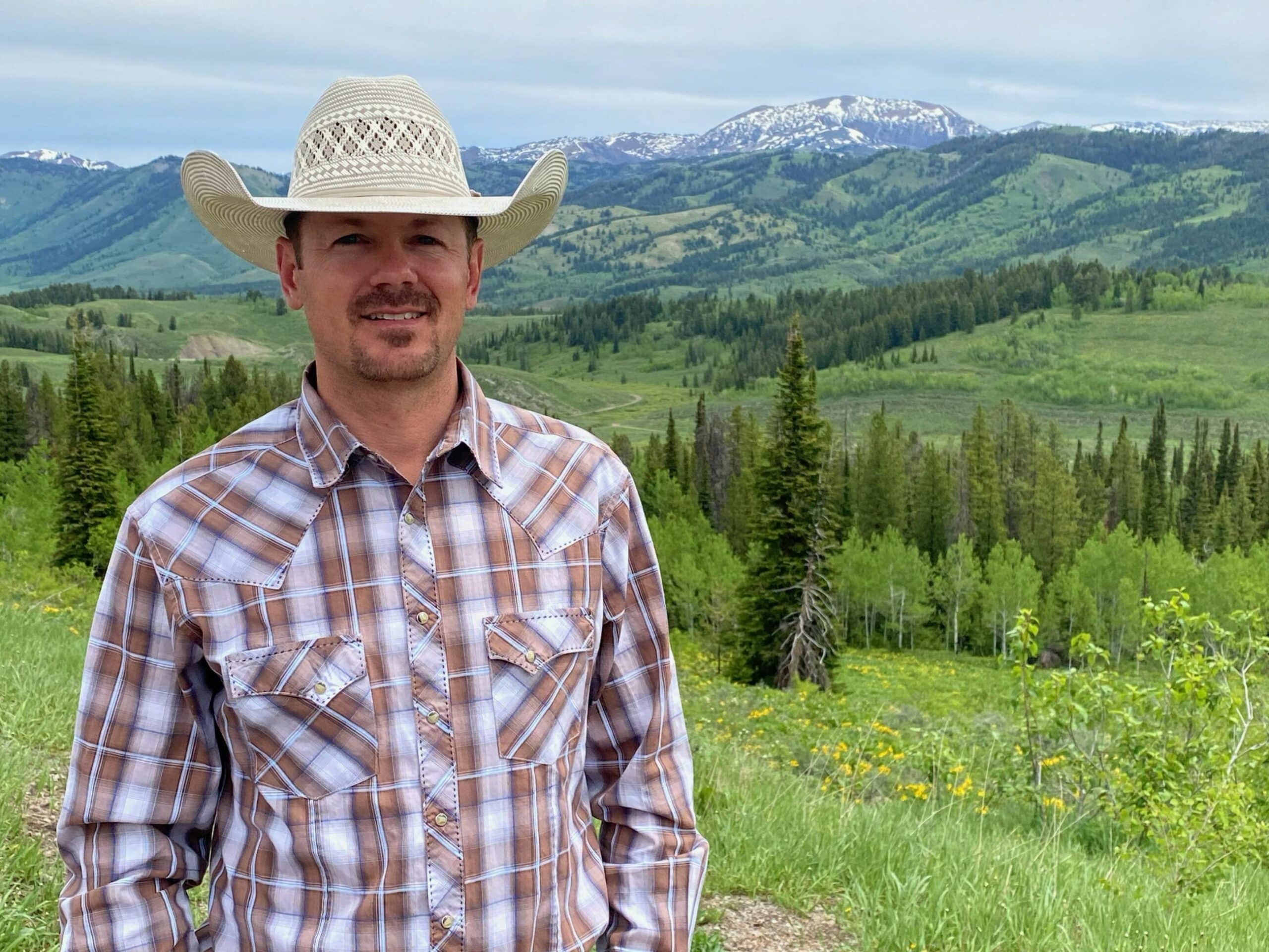 A man in a plaid shirt and cowboy hat stands before a lush green landscape of rolling hills, trees, and distant snow-capped mountains—ideal recreational land or hunting property under a dramatic cloudy sky.