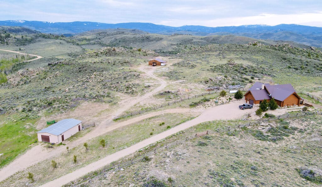 Aerial view of a rural landscape with a log cabin, two outbuildings, dirt roads, and rolling hills in the background under a cloudy sky—ideal as a ranch for sale or hunting property.
