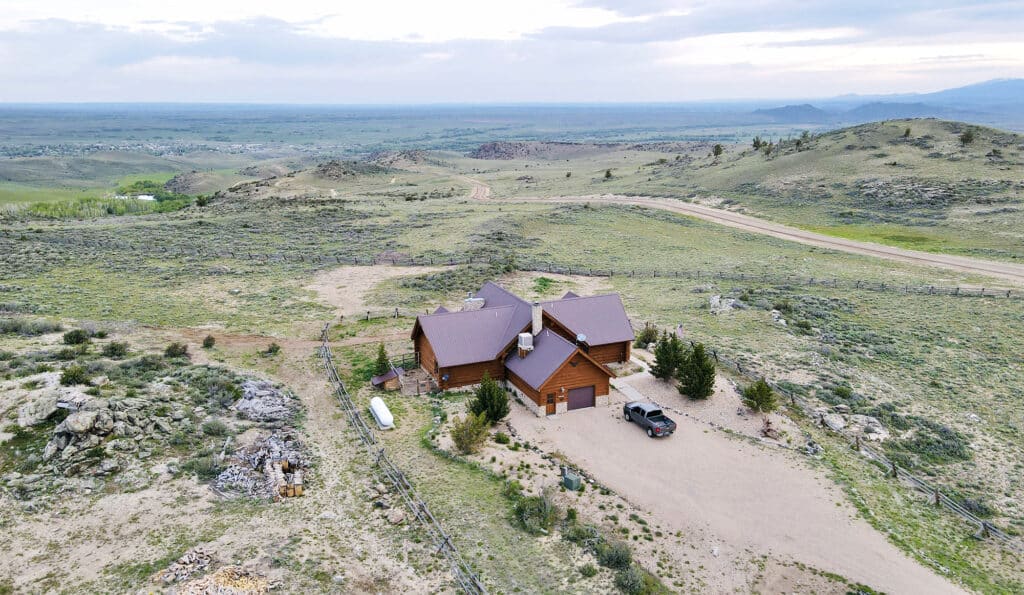 Aerial view of a ranch for sale featuring a wooden house with a brown roof, surrounded by sparse vegetation and hills. The property includes a dirt driveway, parked truck, and expansive hunting land under a partly cloudy sky.