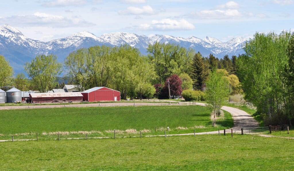 A rural landscape with a red barn and silos, ideal as a cattle ranch or hunting property, surrounded by green fields and trees against snow-capped mountains under a partly cloudy sky.