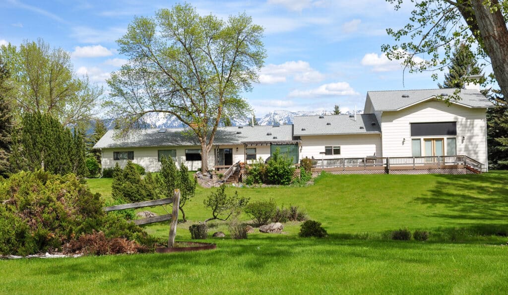A large white house with a gray roof sits on a spacious, well-manicured lawn with trees and shrubs. Mountains and a blue sky with scattered clouds are visible in the background—perfect as a ranch for sale or cattle ranch.