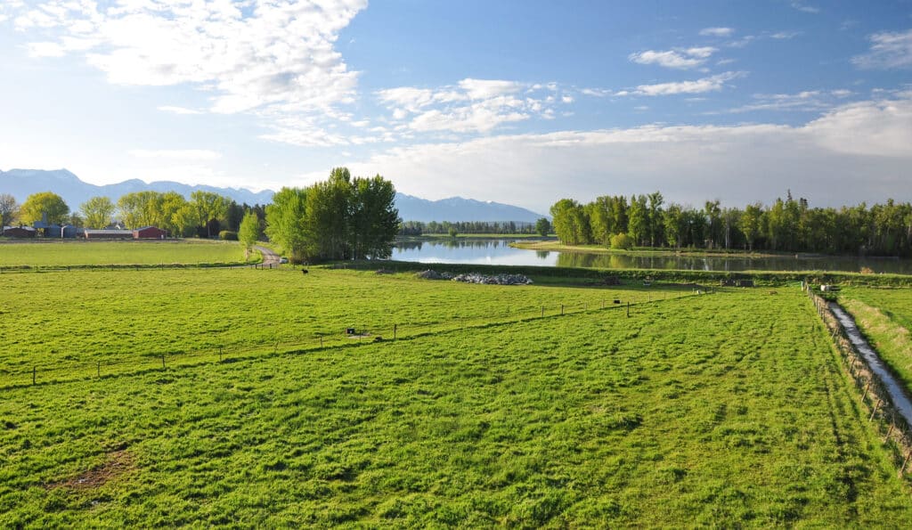 A scenic view of a green pasture on recreational land with a small canal, scattered trees, a pond, and mountains in the background under a partly cloudy blue sky.