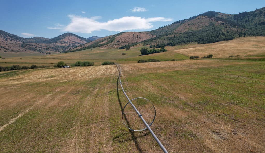 An irrigation pipe with wheels stretches across a dry, grassy field on this cattle ranch, set against rolling hills and mountains beneath a partly cloudy sky. A vehicle is parked off to the left.