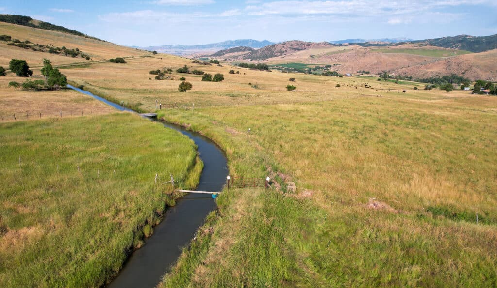 A winding stream flows through a grassy, rolling landscape with hills in the distance. People stand near a small bridge while cattle graze in fields, making this scenic hunting property and cattle ranch land for sale ideal for nature lovers.