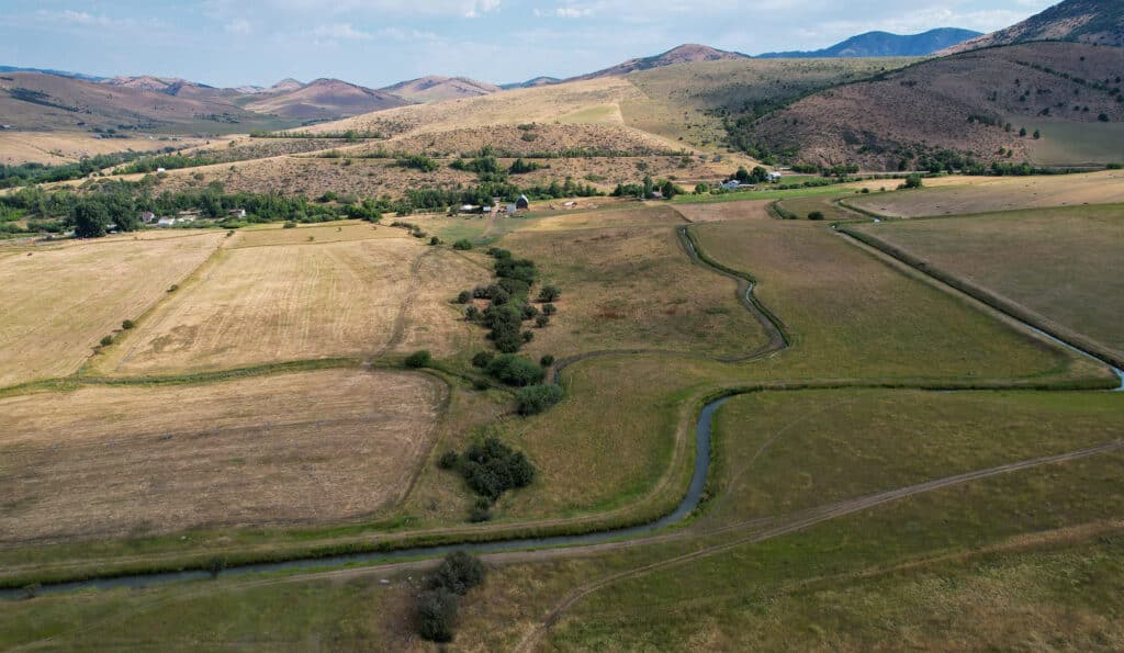 Aerial view of a rural landscape with patchwork fields, a winding stream, green trees, scattered farmhouses, and rolling hills under a mostly clear sky—an ideal hunting property or cattle ranch land for sale.