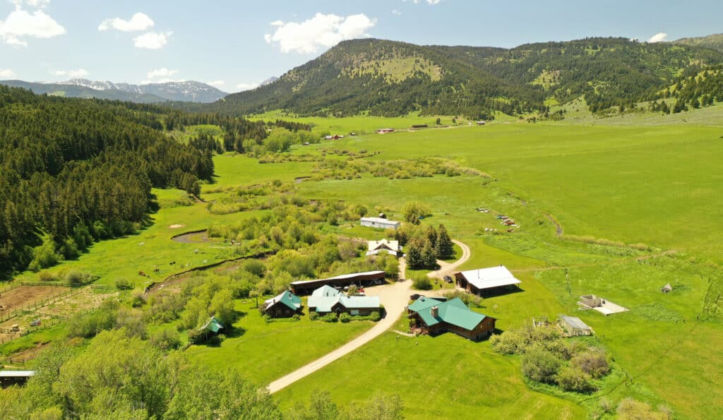 Aerial view of a rural ranch for sale with green fields, scattered buildings with green roofs, winding dirt roads, and forested hills under a blue sky with clouds—an ideal hunting property.