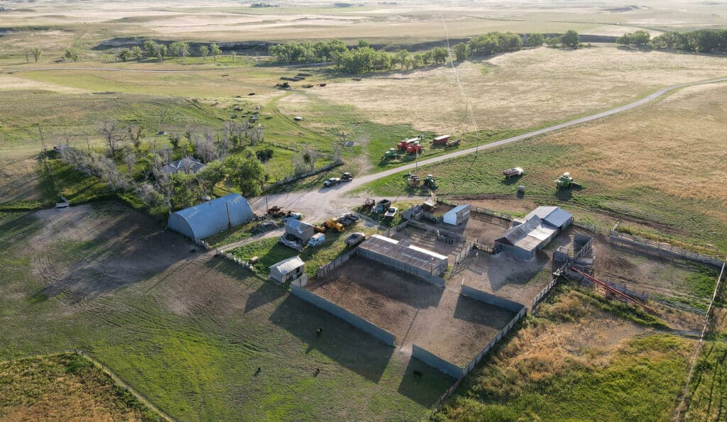 Aerial view of a rural farm with fenced animal pens, barns, vehicles, and equipment, surrounded by open fields and scattered trees under a clear sky—ideal as hunting property or ranch for sale.