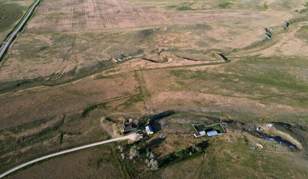 Aerial view of a rural landscape ideal for recreational land or a cattle ranch, with scattered buildings, winding dirt paths, grassy fields, and patches of farmland. A road runs through the bottom left corner near houses and small structures.
