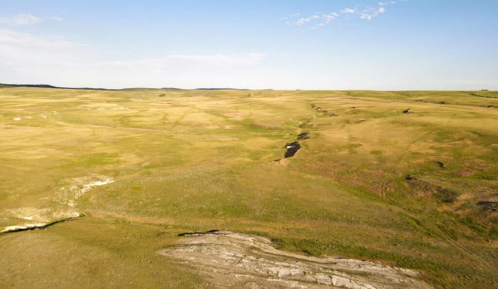 Aerial view of a vast, gently rolling grassland ideal for a cattle ranch or recreational land, with patches of exposed earth and a small ravine running through the landscape under a clear blue sky.