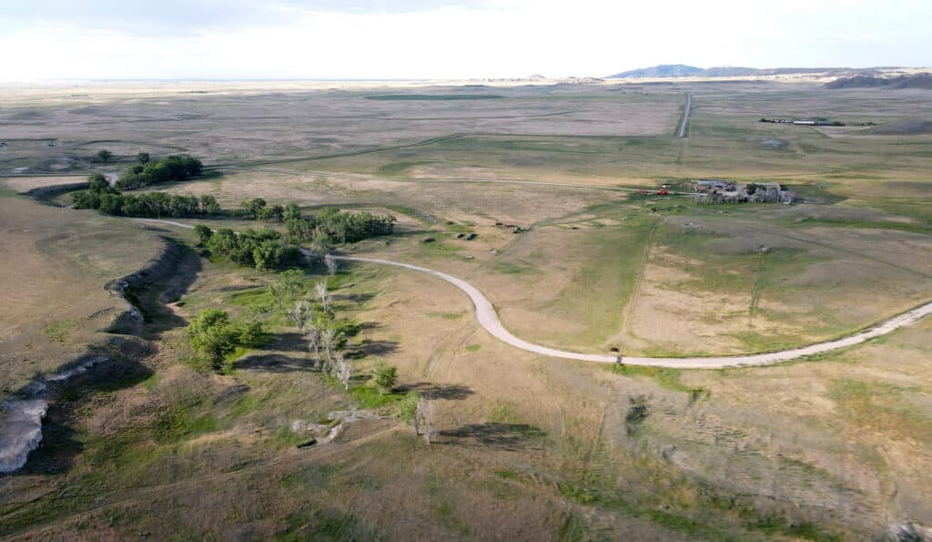 Aerial view of a winding road curving through a vast, open grassland with scattered trees, distant hills, and a few buildings near the road—perfect for a cattle ranch or recreational land under a bright sky.