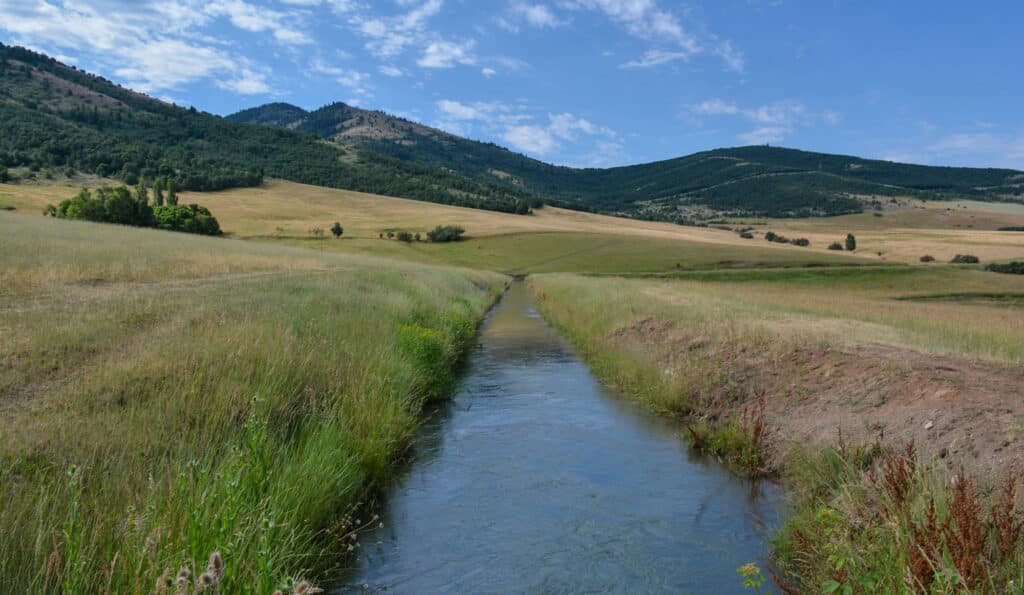 A narrow stream winds through grassy fields on this scenic hunting property, with green rolling hills and mountains in the background under a partly cloudy blue sky.