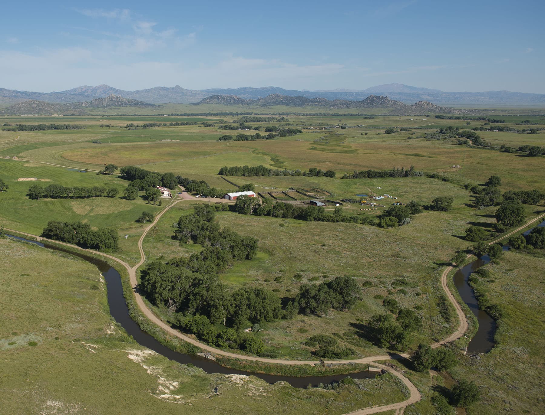 Aerial view of a rural landscape with green fields, a winding creek, farm buildings, and distant hills under a clear blue sky. This picturesque ranch for sale features dirt roads connecting the property and clusters of trees dotting the area.