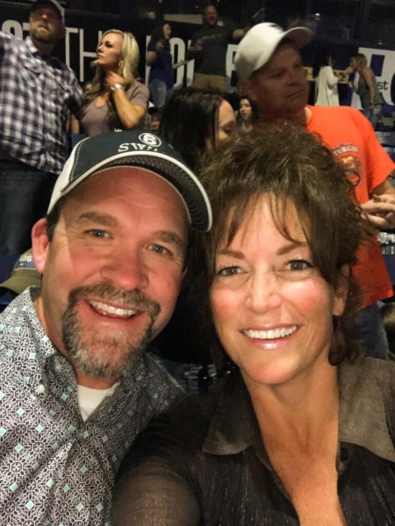 A smiling man in a baseball cap and a woman with wavy hair take a selfie at an indoor event. Several other people, possibly discussing ranch for sale, are visible in the background, some standing and others seated.