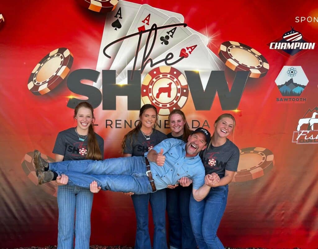 Five people pose smiling in front of a red backdrop featuring playing cards and poker chips. Four women hold up a laughing man horizontally, all in jeans and casual tops. The backdrop says The Show Reno, NV—perfect for fans of ranch for sale adventures.