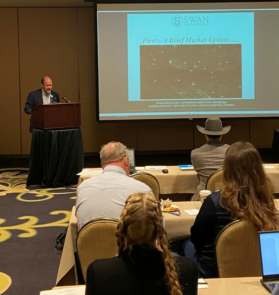 A man stands at a podium giving a presentation on ranch for sale trends to an audience in a conference room. A slide reads First—A Brief Market Update. Attendees sit facing the presenter, some taking notes.