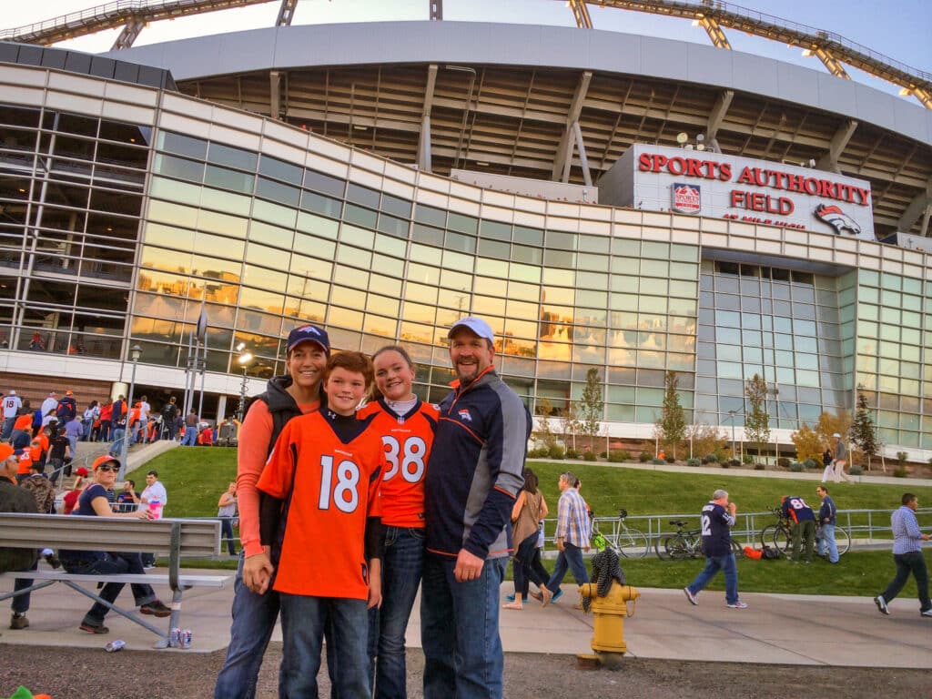 A family of four in Denver Broncos gear smiles outside Sports Authority Field at Mile High stadium, with other fans in the background on a sunny day—perfect for those dreaming of Colorado living or searching for ranch for sale nearby.
