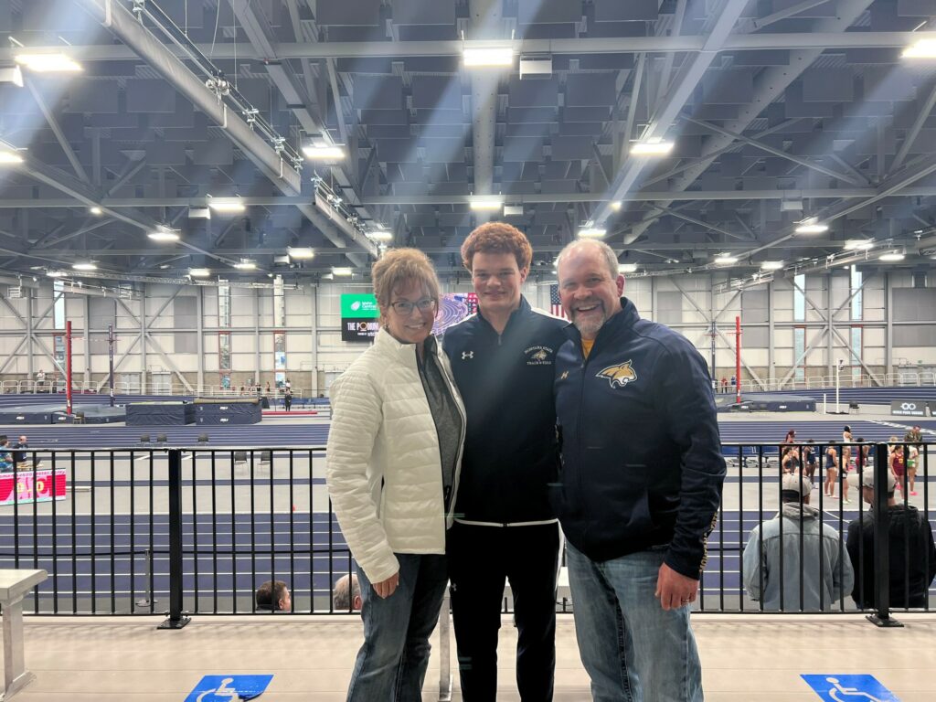 Three people stand smiling in front of an indoor track field. Two adults, one on each side, are wearing jackets, while a young man in athletic apparel stands in the middle. The arena and lanes create a perfect setting to discuss available recreational land.