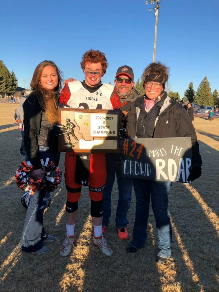 Four people stand on a football field smiling. The player in uniform holds a “2020-2021 5A State Champions” plaque, while another waves a “#24 makes the crowd roar” sign. It's sunny with trees in the background—perfect weather for a hunting property or cattle ranch.