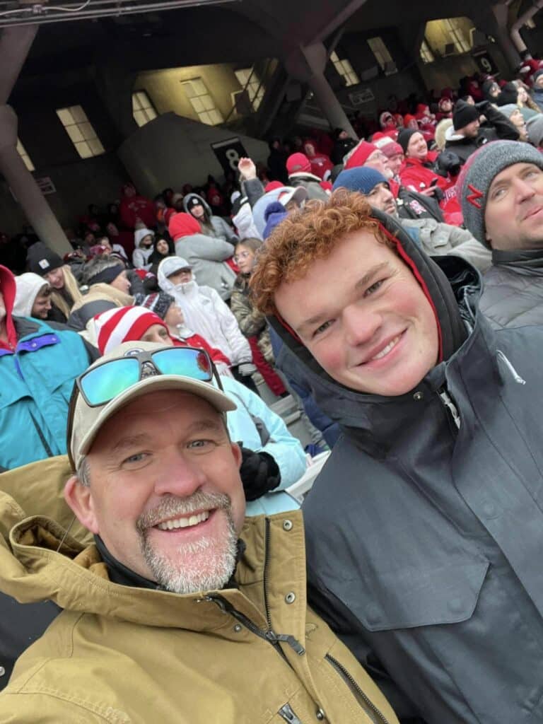 Two people in winter jackets smile for a selfie at an outdoor stadium. Behind them, a crowd of bundled-up spectators enjoys the event, many sporting hats and red attire—perfect for those seeking local fun before browsing ranches or recreational land for sale.