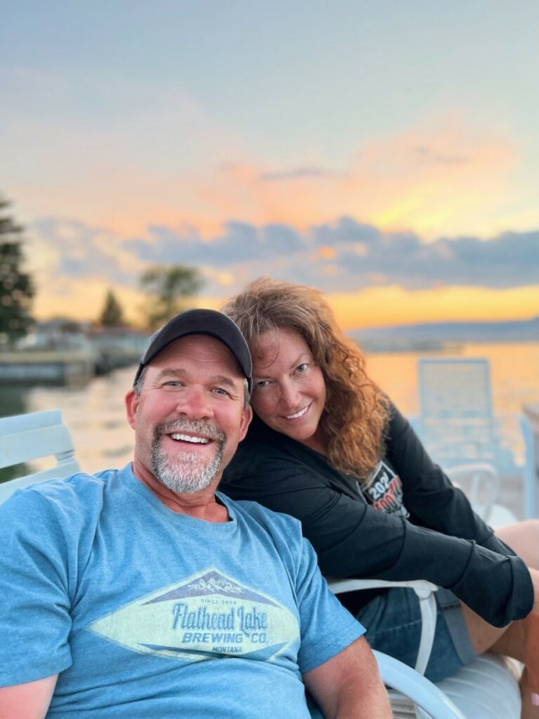 A smiling man in a blue T-shirt and a woman in a black jacket sit close together on outdoor chairs by a lake at sunset, with orange and pink clouds reflecting on the water behind them—perfect for those seeking recreational land or a ranch for sale.