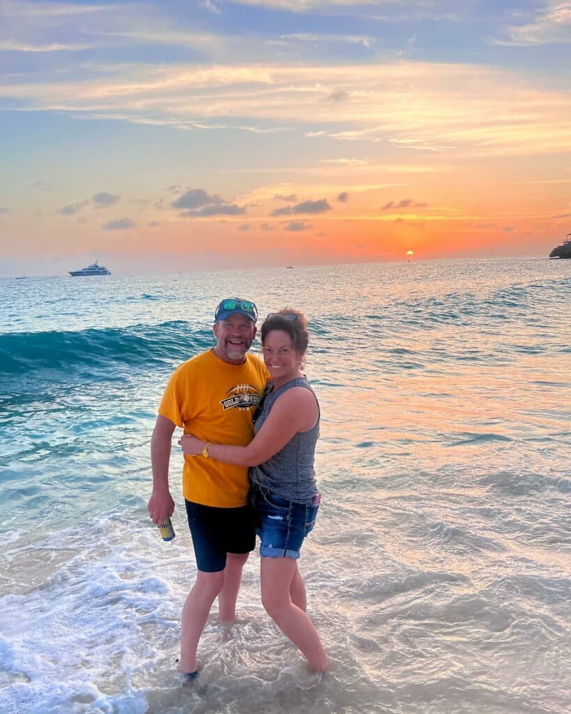 A smiling couple stands in shallow ocean water at sunset, gentle waves around their feet. The man wears a yellow t-shirt and sunglasses; the woman wears a tank top and shorts. A boat drifts in the distance, hinting at endless recreational land adventures.