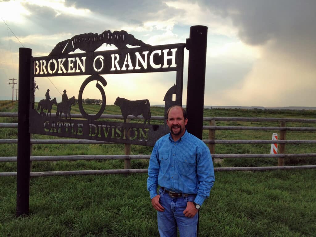 A man in a blue shirt and jeans stands smiling in front of a large “Broken 6 Ranch Cattle Division” sign on a grassy field, showcasing prime cattle ranch and recreational land with a wooden fence and cloudy sky in the background.