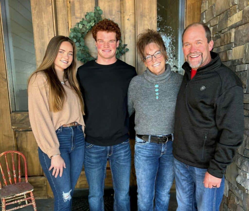 Four people stand smiling in front of a wooden door with a green wreath. Two women and two men, casually dressed in jeans and sweaters, are posed closely together, reflecting the warmth of family found on a ranch for sale.
