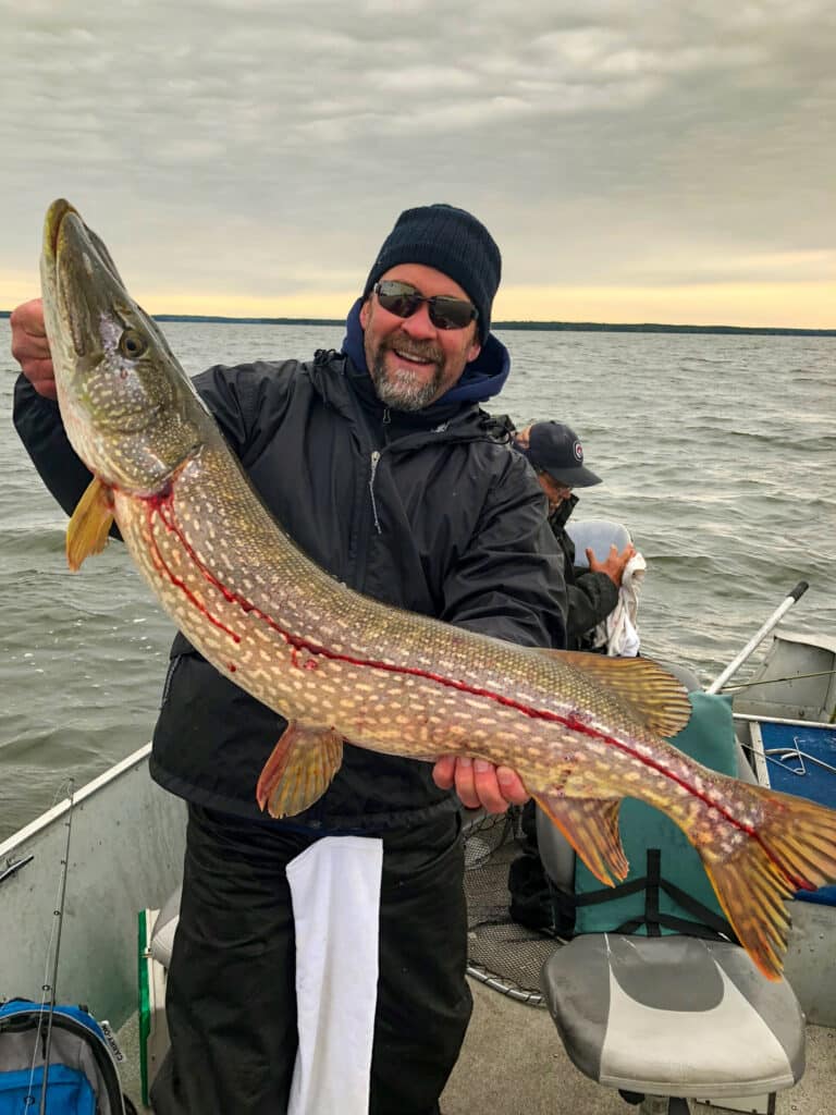 A smiling person wearing sunglasses and a beanie holds a large fish on a boat, enjoying the calm water under cloudy skies. In the background, someone sits with a towel—an ideal day for those seeking recreational land or ranch for sale nearby.