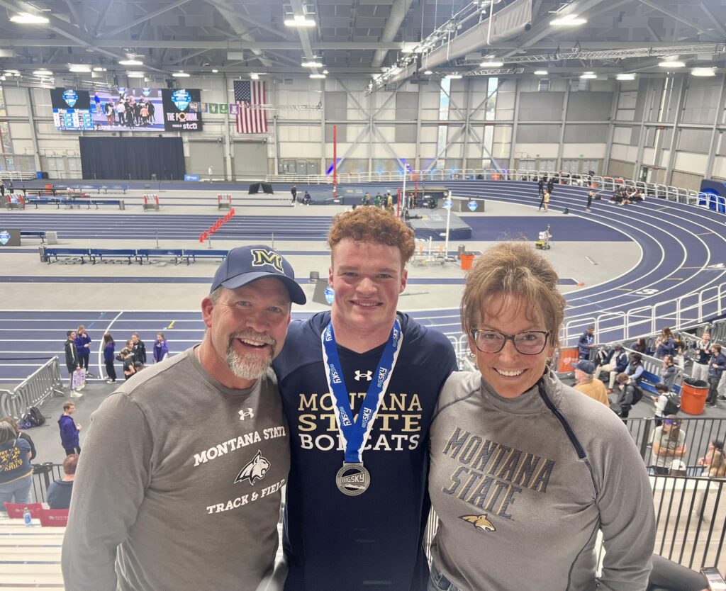 A smiling young athlete wearing a medal stands between two adults, all in Montana State gear, inside an indoor track and field facility with blue lanes—showcasing school pride in the heart of Big Sky Country known for its recreational land.