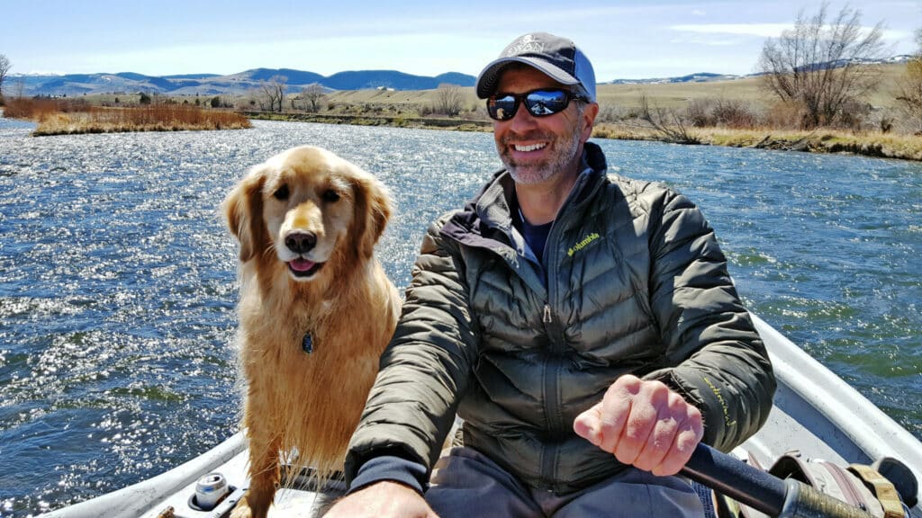 A man wearing sunglasses and a jacket smiles while rowing a boat on a river, with a golden retriever beside him. Grassy hills and mountains of recreational land are visible in the background under a blue sky.