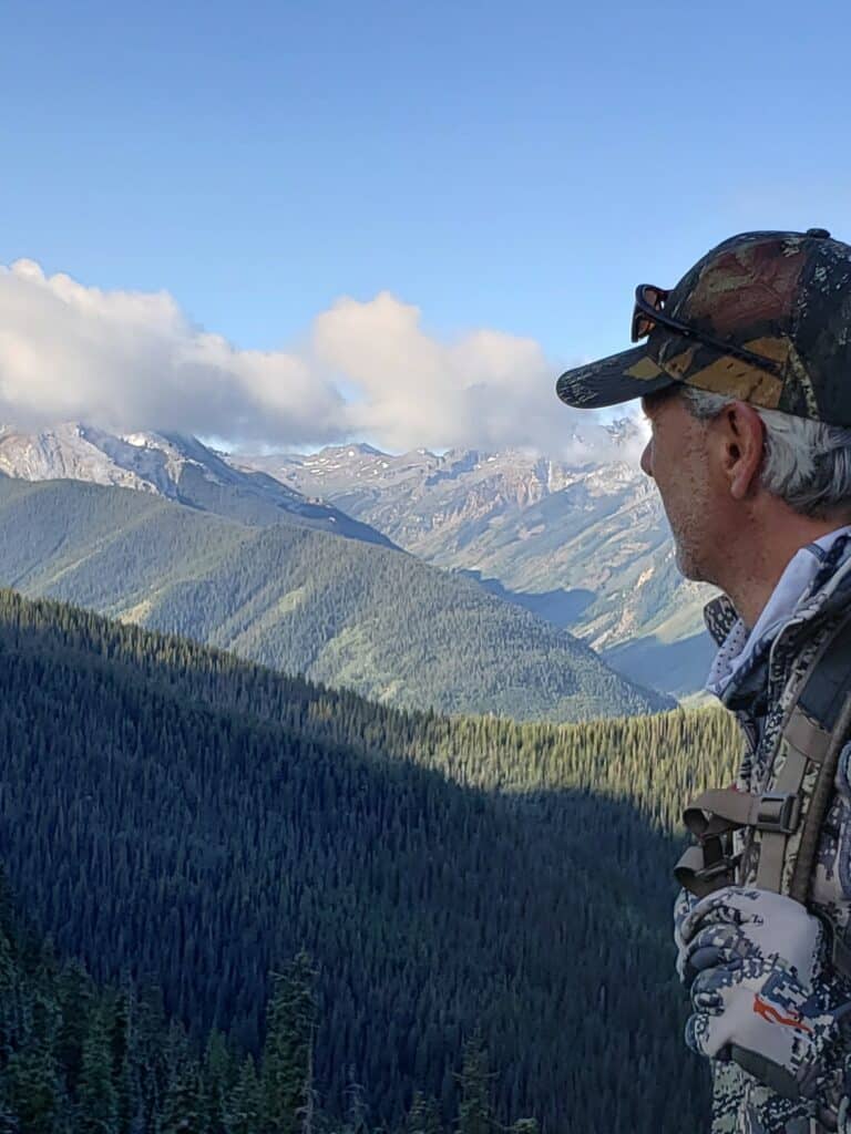 A man in camouflage clothing and hat stands on a mountain, looking out over a forested valley with distant snow-capped peaks under a blue sky—prime recreational land near a ranch for sale.