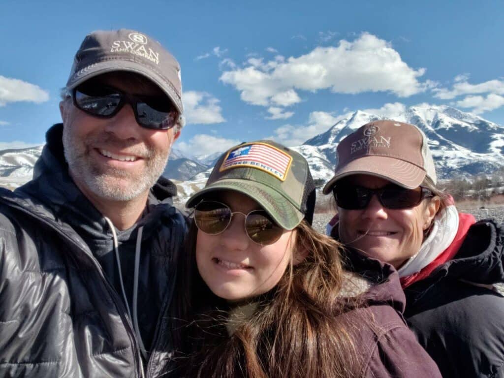 Three people in sunglasses and hats pose for a selfie outdoors, with snowy mountains and a partly cloudy sky behind them. Bundled in jackets, they smile at the camera, enjoying the beauty of this recreational land near a scenic cattle ranch.