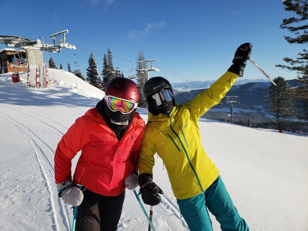 Two people in bright ski jackets and helmets pose cheerfully on a snowy ski slope, with ski poles in hand and rugged mountains—perfect for hunting property or ranch for sale—plus trees and a ski lift visible under a clear blue sky.