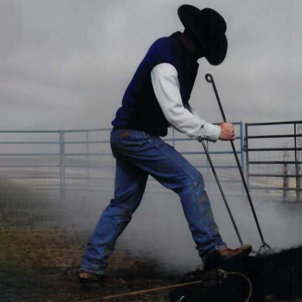 A person in a cowboy hat, jeans, and boots stands on smoky ground outdoors near a fenced area, working with long metal rods—capturing the authentic atmosphere of life on a cattle ranch.