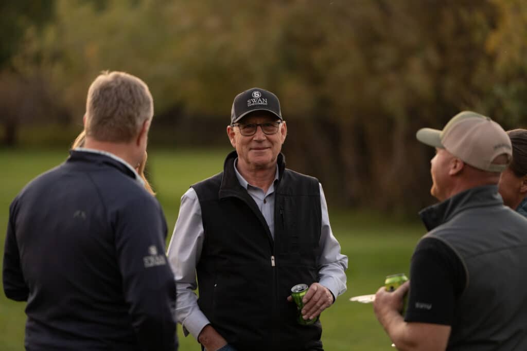 A group of men are standing outdoors on grass, talking and holding drinks. One man in the center wears glasses, a black vest, and a cap, smiling at the camera. Trees are blurred in the background on this beautiful cattle ranch.