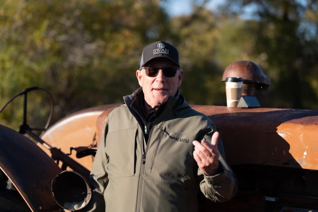 A man in sunglasses and a cap stands outdoors in front of a rusty vehicle, gesturing with one hand. He wears a green jacket, with a coffee cup on the vehicle behind him—an ideal setting for showcasing recreational land or cattle ranch opportunities.