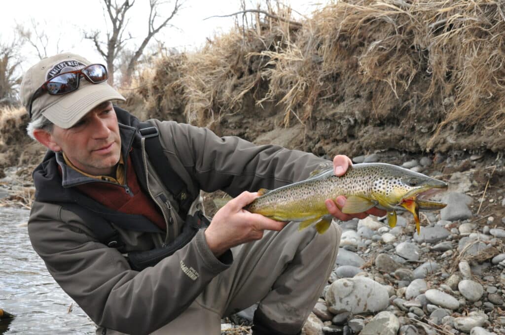 A man kneels by a rocky stream bank, holding a large brown trout with both hands. Wearing outdoor gear, he enjoys the outdoors on recreational land. Dry grass and bare trees are visible in the background.
