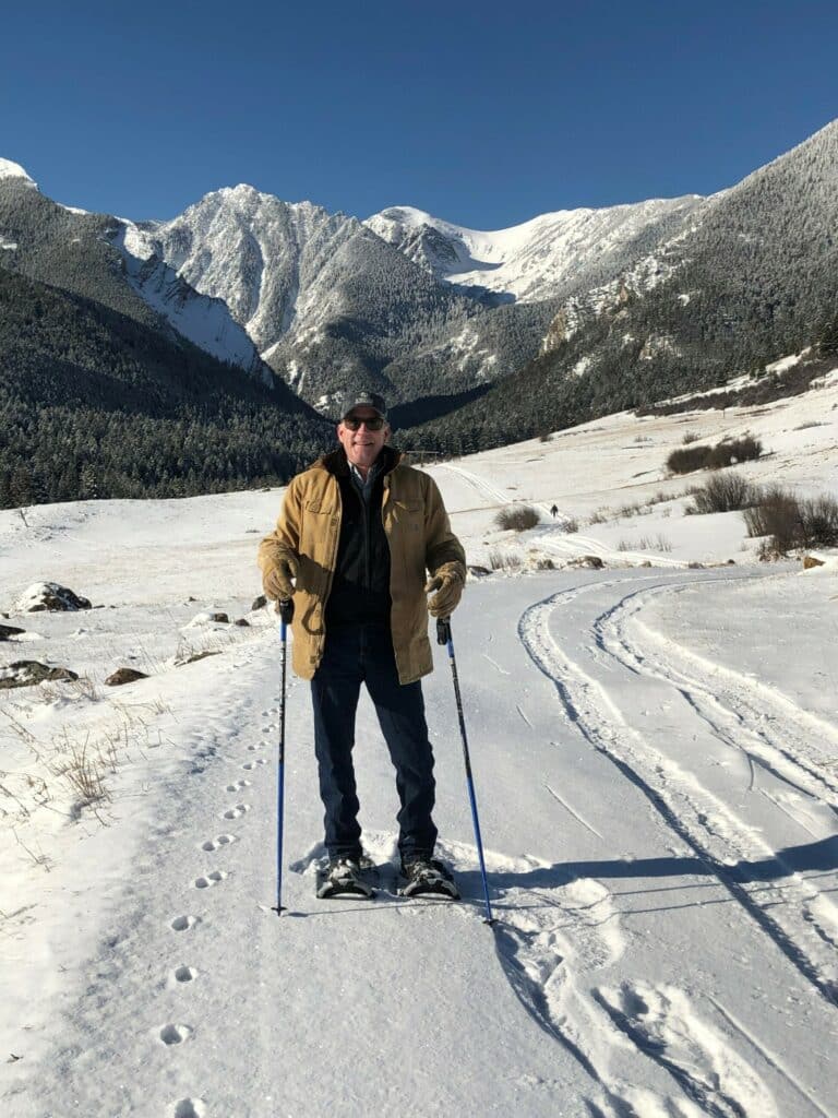 A person wearing a tan jacket and black pants is snowshoeing on a snowy path surrounded by mountains and trees under a clear blue sky, exploring scenic recreational land.