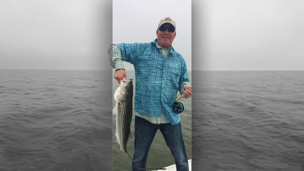 A man wearing sunglasses and a blue plaid shirt stands on a boat holding a large fish in one hand and a fishing reel in the other, with recreational land and open water beneath a cloudy sky in the background.