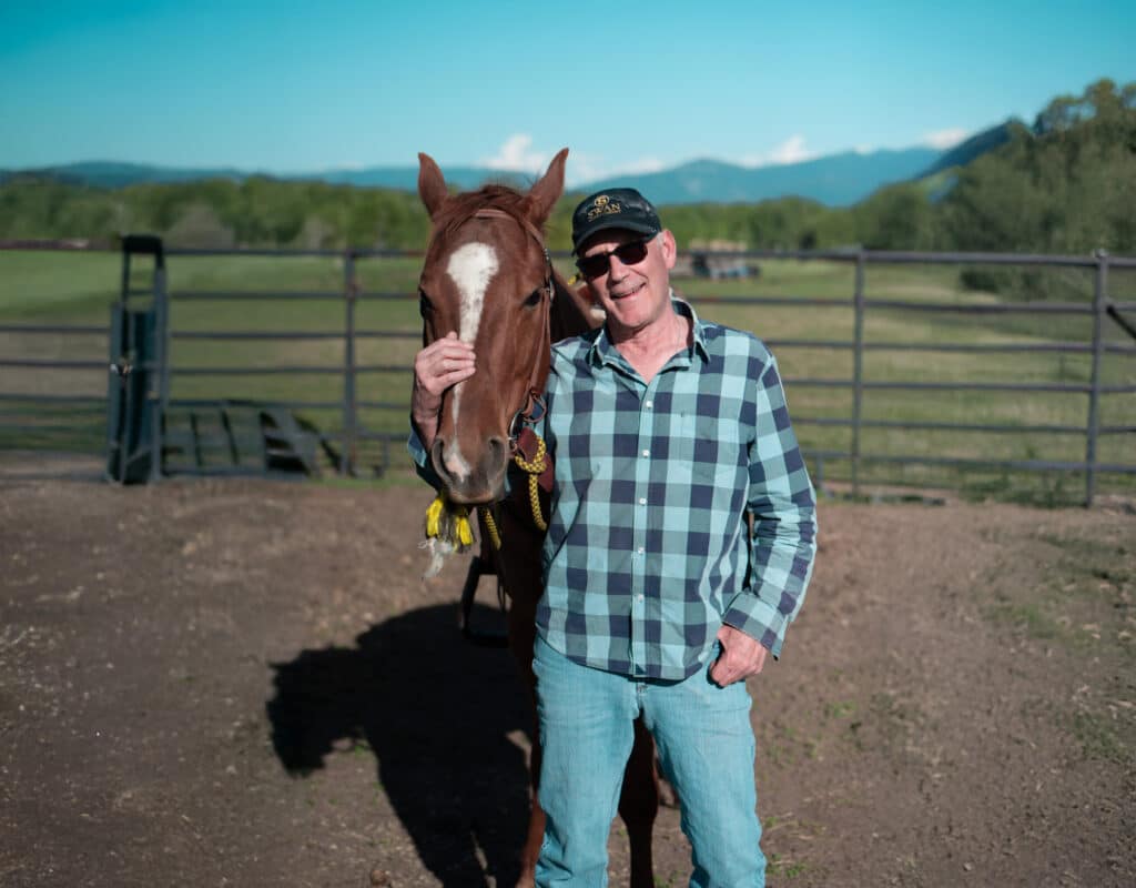A man in a plaid shirt and cap stands next to a brown horse, smiling with one hand on the horse’s face. They are outdoors in a fenced area, perfect for a cattle ranch or hunting property, with green fields and mountains in the background.