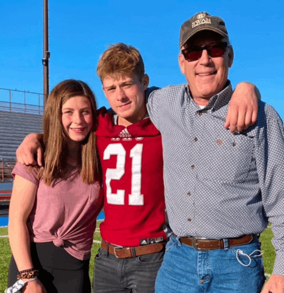 Three people stand together on recreational land outdoors on a sunny day. A young woman is on the left, a young man in a red football jersey (#21) is in the middle, and an older man in sunglasses and a cap is on the right, all smiling.