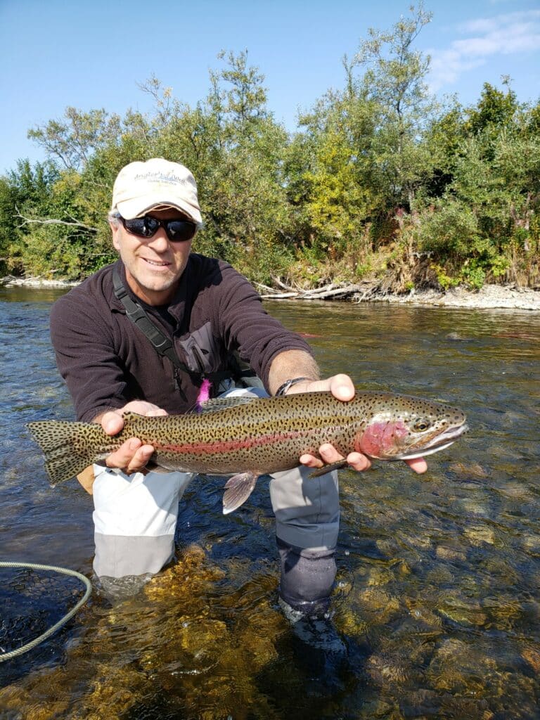 A man wearing sunglasses and a cap stands in a shallow stream on hunting property, holding a large rainbow trout with both hands. Green trees and blue sky fill the background.