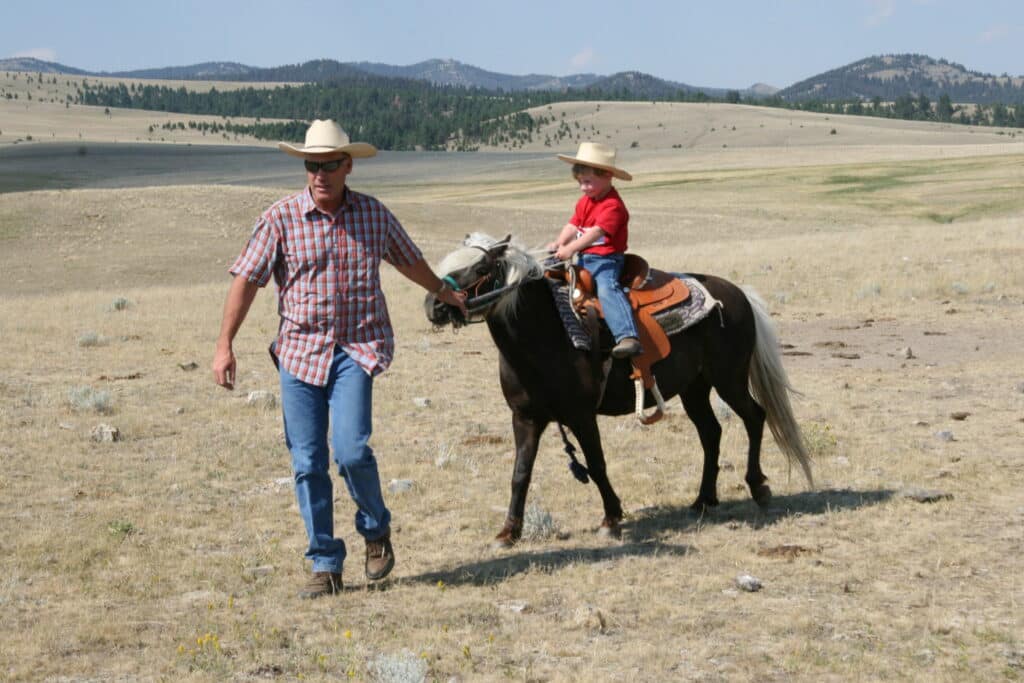 A man in a cowboy hat leads a small horse with a young child, also in a hat, riding on it across dry, grassy recreational land with mountains in the background—perfect for those seeking cattle ranch opportunities.