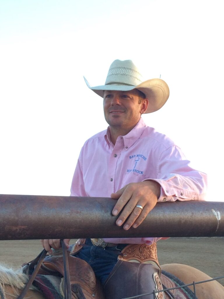 A man in a white cowboy hat and pink shirt sits on a horse, leaning on a metal fence. He is outdoors under a clear sky, with cattle ranch land for sale visible in the scenic background.