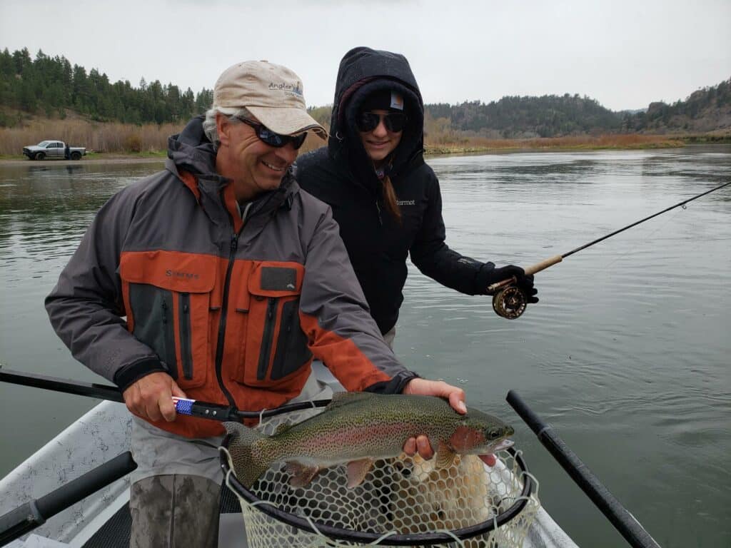 Two people in jackets and sunglasses stand in a boat on a river. One holds a large rainbow trout in a net, while the other holds a fishing rod. Trees and hills, ideal for a hunting property or cattle ranch, are visible in the background.