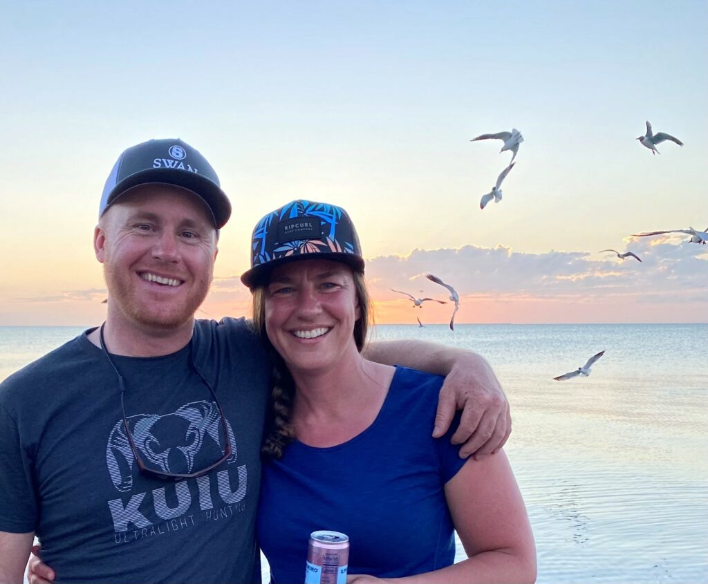 A smiling man and woman stand arm-in-arm on a beach at sunset, with seagulls flying over the calm water behind them. Both wear casual clothing and hats, enjoying their time together while dreaming of hunting property by the shore.
