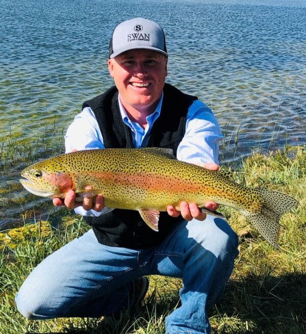 A smiling man in a cap and vest kneels by a lake on recreational land, holding a large, colorful trout with both hands. The water and grassy shoreline are visible in the background.