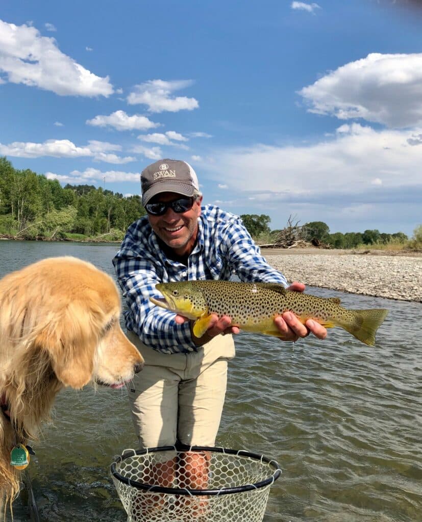 A smiling man in sunglasses and a cap holds a brown trout over a river on prime hunting property, while a golden retriever looks at the fish beside a fishing net in shallow water. Trees and a rocky bank complete the scenic background.
